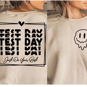 以下が含まれることがあります： 背面に「Test Day」が複数回と「Just Do Your Best」と書かれた黒いグラフィックが施されたベージュのトレーナー。トレーナーの前面には、溶けているスマイリーフェイスの黒いグラフィックがあります。