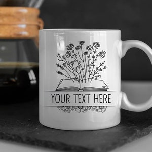 May include: White ceramic mug with a black line drawing of an open book with flowers growing out of it. The text "YOUR TEXT HERE" is printed on the mug.