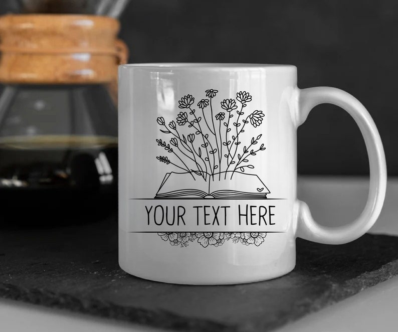 May include: White ceramic mug with a black line drawing of an open book with flowers growing out of it. The text "YOUR TEXT HERE" is printed on the mug.