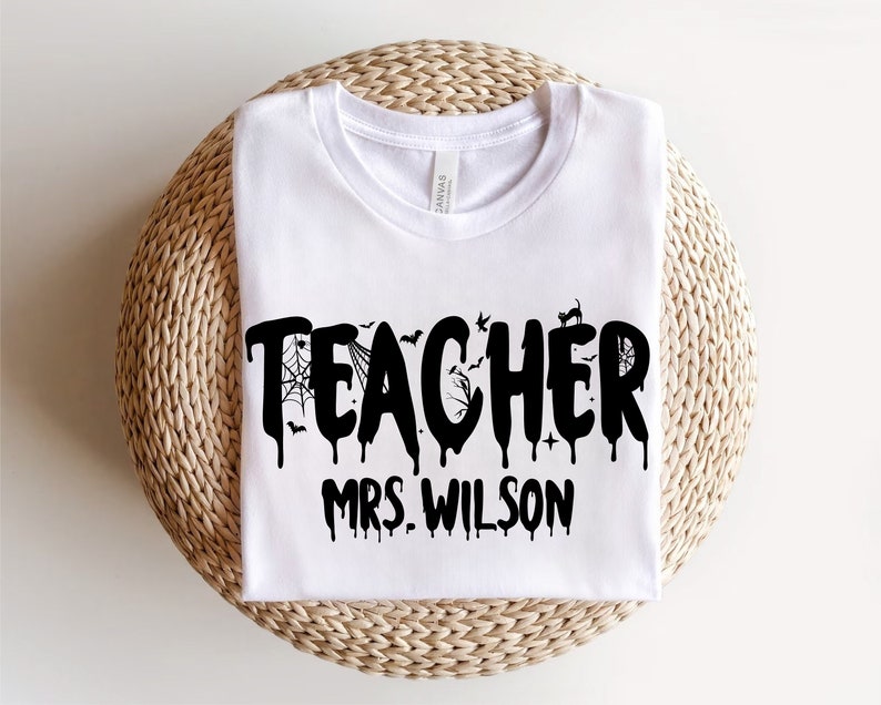 May include: White t-shirt featuring the word "TEACHER" in a black, dripping font, with Halloween-themed decorations. The name "MRS. WILSON" is printed underneath. The shirt is folded and placed on a woven, circular surface.