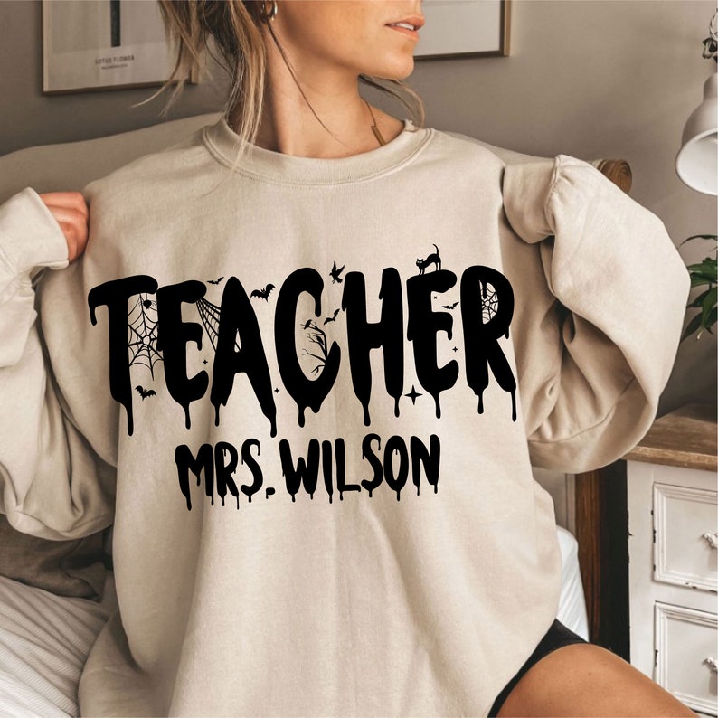 May include: A beige sweatshirt featuring the word "TEACHER" in large, dripping black letters, adorned with bats, a spiderweb, and a black cat. The name "MRS. WILSON" is printed below in black. The sweatshirt is a relaxed, comfortable style.
