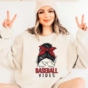 May include: A white sweatshirt with a graphic of a woman with a red bandana in her hair, wearing sunglasses with baseballs on the lenses. The text "BASEBALL VIBES" is printed below the graphic.