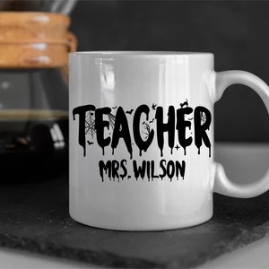 May include: White ceramic mug with the word "TEACHER" in black, dripping font, decorated with bats, a spiderweb, and a black cat. The name "MRS. WILSON" is printed below. The mug is on a dark surface.