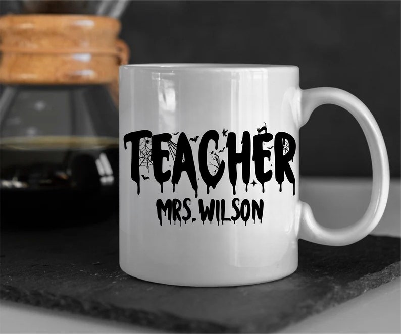 May include: White ceramic mug with the word "TEACHER" in black, dripping font, decorated with bats, a spiderweb, and a black cat. The name "MRS. WILSON" is printed below. The mug is on a dark surface.