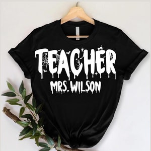 May include: Black t-shirt featuring the word "TEACHER" in dripping white letters, and "MRS. WILSON". The design incorporates bats and a spider web, hinting at a Halloween theme. The shirt is displayed on a wooden hanger.