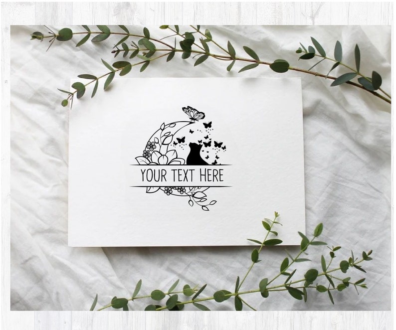 May include: Black and white illustration of a cat sitting in a circle of flowers and butterflies. The text "YOUR TEXT HERE" is written on a banner below the cat.