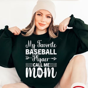 May include: A dark green sweatshirt with white text that reads "My Favorite Baseball Player Call Me Mom" with a baseball and arrow graphic.
