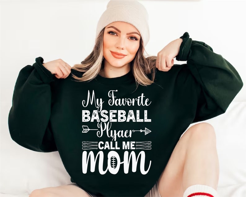 May include: A dark green sweatshirt with white text that reads "My Favorite Baseball Player Call Me Mom" with a baseball and arrow graphic.