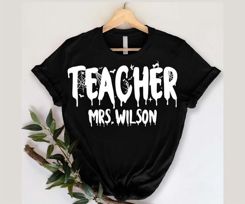 May include: Black t-shirt featuring the word "TEACHER" in dripping white lettering, with "MRS. WILSON" below. The design incorporates bats and a spiderweb, hinting at a Halloween theme. The shirt is displayed on a wooden hanger.