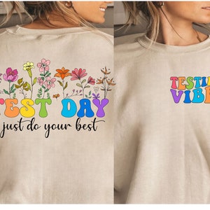 Peut inclure: Un sweat-shirt beige clair avec un motif floral arc-en-ciel et le texte "TEST DAY just do your best" au dos. Le devant du sweat-shirt porte le texte "TESTING VIBES" en lettres arc-en-ciel.