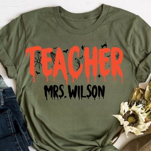 May include: An olive green t-shirt featuring the word "TEACHER" in dripping orange lettering, with bat and spiderweb motifs. The name "MRS. WILSON" is printed below in black. A Halloween-themed garment.