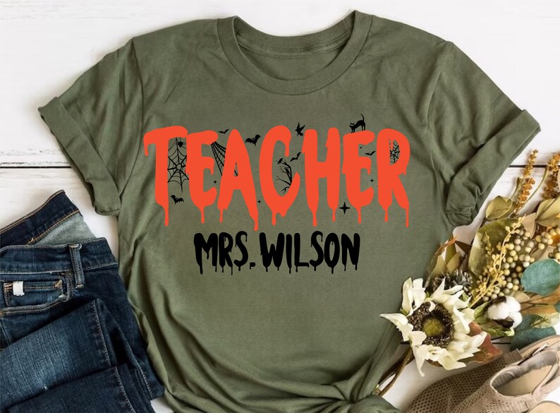 May include: An olive green t-shirt featuring the word "TEACHER" in dripping orange lettering, with bat and spiderweb motifs. The name "MRS. WILSON" is printed below in black. A Halloween-themed garment.