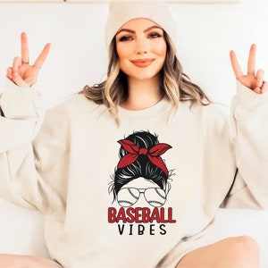 May include: A white sweatshirt with a graphic of a woman with a red bandana in her hair, wearing sunglasses with baseballs on the lenses. The text "BASEBALL VIBES" is printed below the graphic.
