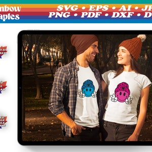 Puede incluir: Una pareja vistiendo camisetas blancas con un diseño gráfico de cara sonriente. El diseño gráfico presenta un gorro azul y rosa con un corazón y un signo de paz. La pareja está de pie en un parque con árboles y follaje de otoño en el fondo.