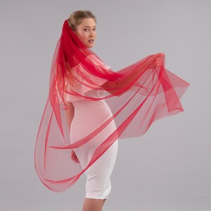 May include: A single-layer red tulle wedding veil. The veil is draped over the shoulders and held in one hand.