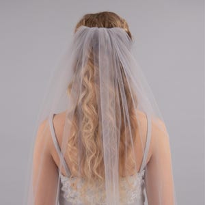 Op de afbeelding: Een eenlaags bruids sluier van wit tule met een gegolfde rand. De sluier wordt gedragen door een persoon met lang blond haar.