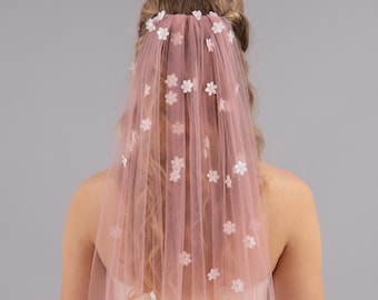 May include: A blush pink tulle veil decorated with white flower embellishments. The veil is attached at the crown and falls down the back. The small flowers are scattered across the veil, creating a soft, romantic appearance.