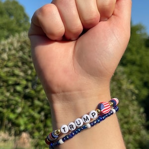K&ouml;nnte beinhalten: Ein Perlenarmband mit dem Wort "TRUMP" in wei&szlig;en Perlen mit schwarzer Beschriftung. Das Armband hat au&szlig;erdem einen kleinen amerikanischen Flaggenanh&auml;nger und blaue und wei&szlig;e Perlen.