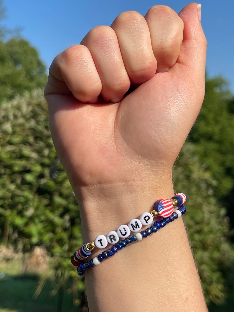 K&ouml;nnte beinhalten: Ein Perlenarmband mit dem Wort "TRUMP" in wei&szlig;en Perlen mit schwarzer Beschriftung. Das Armband hat au&szlig;erdem einen kleinen amerikanischen Flaggenanh&auml;nger und blaue und wei&szlig;e Perlen.