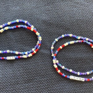 K&ouml;nnte beinhalten: Zwei Perlenarmb&auml;nder mit roten, wei&szlig;en und blauen Perlen. Ein Armband tr&auml;gt den Schriftzug "USA", das andere den Schriftzug "TRUMP".