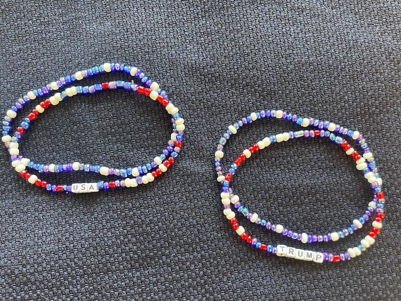 K&ouml;nnte beinhalten: Zwei Perlenarmb&auml;nder mit roten, wei&szlig;en und blauen Perlen. Ein Armband tr&auml;gt den Schriftzug "USA", das andere den Schriftzug "TRUMP".
