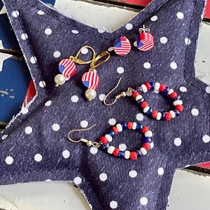 K&ouml;nnte beinhalten: Ein Set aus drei patriotischen Ohrringen in den Farben Rot, Wei&szlig; und Blau. Die Ohrringe werden auf einem blauen Stoff mit wei&szlig;en Punkten pr&auml;sentiert. Ein Paar Ohrringe hat einen kleinen amerikanischen Flaggen-Anh&auml;nger, w&auml;hrend das andere Paar einen Perlenkreis mit roten, wei&szlig;en und blauen Perlen hat.