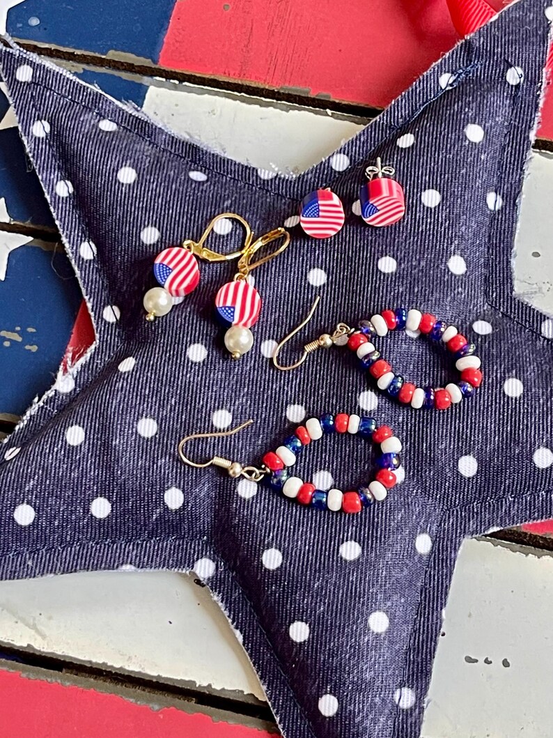 K&ouml;nnte beinhalten: Ein Set aus drei patriotischen Ohrringen in den Farben Rot, Wei&szlig; und Blau. Die Ohrringe werden auf einem blauen Stoff mit wei&szlig;en Punkten pr&auml;sentiert. Ein Paar Ohrringe hat einen kleinen amerikanischen Flaggen-Anh&auml;nger, w&auml;hrend das andere Paar einen Perlenkreis mit roten, wei&szlig;en und blauen Perlen hat.