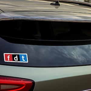 May include: A rear view of a silver car with a black and white sticker on the back window. The sticker says "fdt" in white letters with a red "f" and a blue "t".