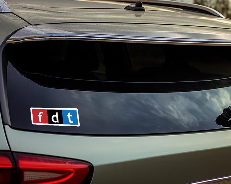 May include: A rear view of a silver car with a black and white sticker on the back window. The sticker says "fdt" in white letters with a red "f" and a blue "t".