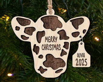 May include: A wooden cow-shaped Christmas ornament with a brown and white cow pattern. The ornament has the text "Merry Christmas" and a tag with the text "Olivia 2025".