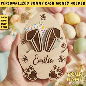 Op de afbeelding: Een houten paasei-vormige geldhouder met konijnenoren en pootafdrukken. De houder is gepersonaliseerd met de naam "Emilia" en bevat contant geld en kleurrijke kaarten. De tekst "PERSONALIZED BUNNY CASH MONEY HOLDER" staat bovenaan.