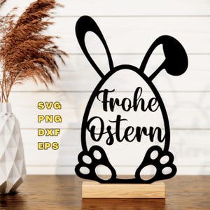 May include: Black silhouette of a bunny rabbit holding an egg with the text "Frohe Ostern" on it. The bunny is standing on a wooden base.