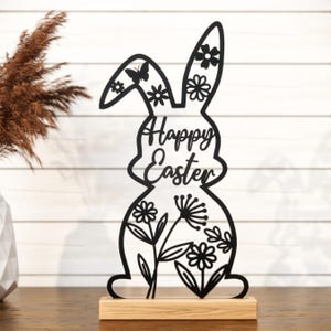 May include: Black metal cutout of a bunny rabbit with floral designs and the text "Happy Easter". The bunny is standing on a wooden base.