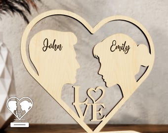 May include: A wooden heart-shaped silhouette of a couple with the names "John" and "Emily" engraved on it. The word "LOVE" is cut out of the heart with a small heart in the middle of the "O". The heart is on a wooden stand.