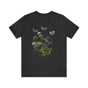 May include: Dark heather gray t-shirt featuring a black raven perched among green roses and leaves. Three butterflies and a moth are also depicted. The design is a grayscale illustration with green accents.