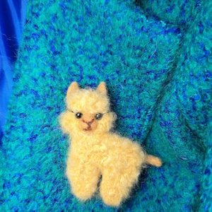 以下が含まれることがあります： 黒いワイヤーフレームのメガネをかけた、小さな黄色のアルパカのピン。アルパカはふわふわした質感で、小さな尻尾があります。ピンは、テクスチャ加工されたターコイズと青の背景に配置されています。