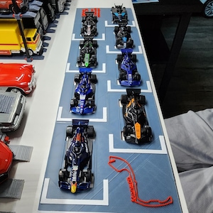 May include: A collection of miniature Formula 1 race cars in various colors, including blue, black, and green, are arranged on a blue surface with white lane markings. A red outline of a race track is also present. The cars are detailed and appear to be model replicas.