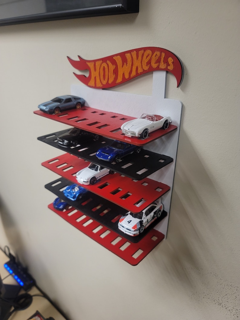 May include: A red and black wooden Hot Wheels display shelf with a red and orange Hot Wheels logo. The shelf has five levels and each level has slots for displaying toy cars. There are five toy cars displayed on the shelf.