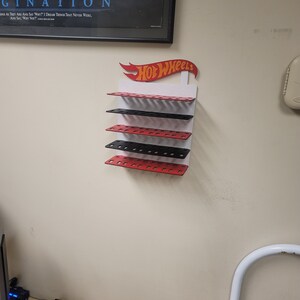May include: A white wall mounted display rack with five shelves for storing Hot Wheels toy cars. The shelves are alternating red and black. The Hot Wheels logo is red and yellow with a red flame design.