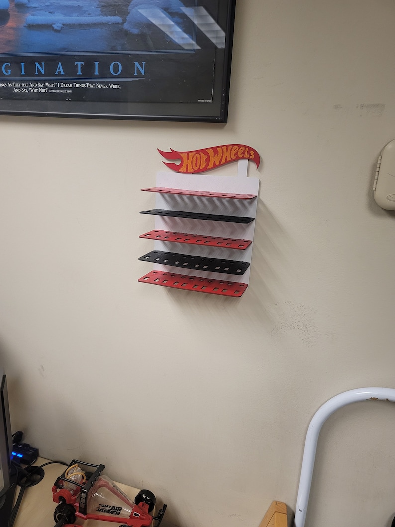 May include: A white wall mounted display rack with five shelves for storing Hot Wheels toy cars. The shelves are alternating red and black. The Hot Wheels logo is red and yellow with a red flame design.