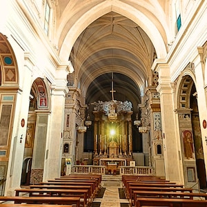 May include: Interior view of a church with arched ceilings and rows of wooden pews. The altar is at the end of the nave, illuminated by a bright light. The walls are adorned with paintings and decorative elements.