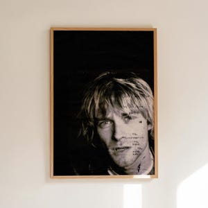 May include: Black and white framed portrait of a man with light-colored hair. The image has text overlayed on the face. The frame is a light brown color. The background is black.