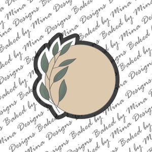 May include: A beige circle with a black outline and a leafy branch design in shades of green. The circle is on a white background with the text "Baked by Mina Designs" repeated.