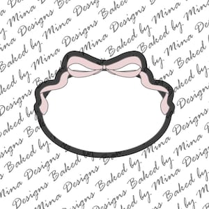 May include: A cookie cutter in the shape of a decorative label with a bow. The cutter has a black outline with a light pink bow and ribbon detail. The background has a repeating pattern of text that reads "Baked by Mina Designs".