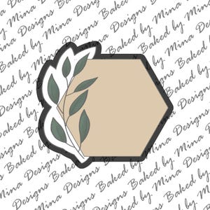 Puede incluir: Un cortador de galletas hexagonal con un diseño de rama de hojas. El cortador es beige con un contorno gris oscuro y las hojas son verdes.