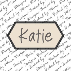 May include: A beige hexagonal sign with a black border and the name "Katie" written in black cursive lettering.