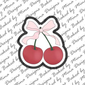 May include: A cherry-shaped cookie cutter featuring two red cherries with green stems and a light pink bow. The cutter has a black outline and is designed for baking. The background has a repeating pattern of text.