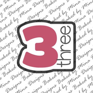 Può includere: Un tagliapasta a forma di numero 3 con la parola "three" scritta verticalmente. Il numero è di colore rosa intenso, bordato di nero, su sfondo bianco. Il testo "Baked by Mina Designs" è ripetuto in diagonale.