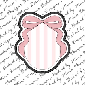 May include: A cookie cutter with a black outline shaped like an oval plaque featuring a large pink bow at the top. The center of the cutter has a white and light pink vertical striped pattern. The design is centered on a white background.