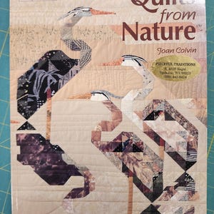 May include: A softcover book titled Quilts from Nature by Joan Colvin, featuring a quilted cover design of three herons in shades of beige, brown, and black. The cover includes the text That Patchwork Place and a Pieceful Traditions sticker.